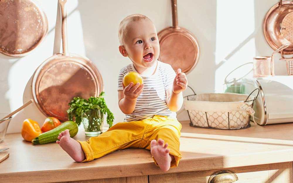 Baby sitzt lachend auf der Küchentheke und hält eine Zitrone in der Hand
