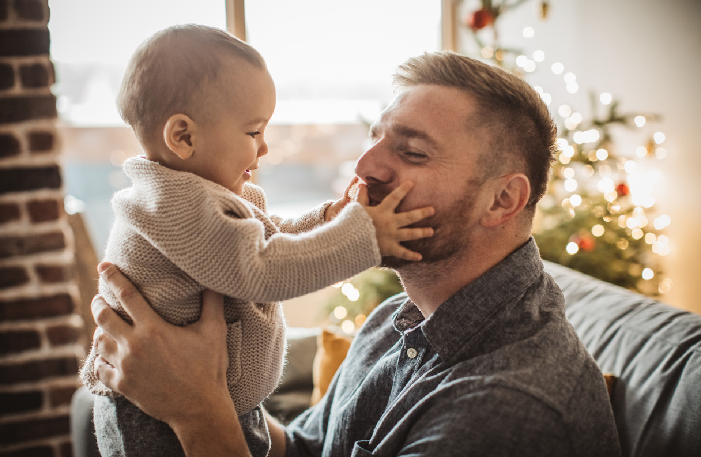 Vater auf der Couch in weihnachtlich geschmücktem Wohnzimmer mit Baby auf dem Arm