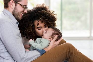 Glücklicher Vater hält sein Baby auf demScho, die Mutter küsst es zärtlich auf die Wange