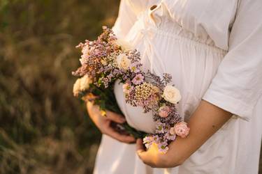 Bildausschnitt: Hochschwangere Frau in weißem Kleid hält Blumenkranz vor ihrem Bauch