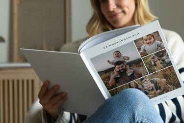 Junge Frau blättert lächelnd in einem Fotobuch mit Hardcovereinband, das Cover zeigt eine Collage mit Familienfotos